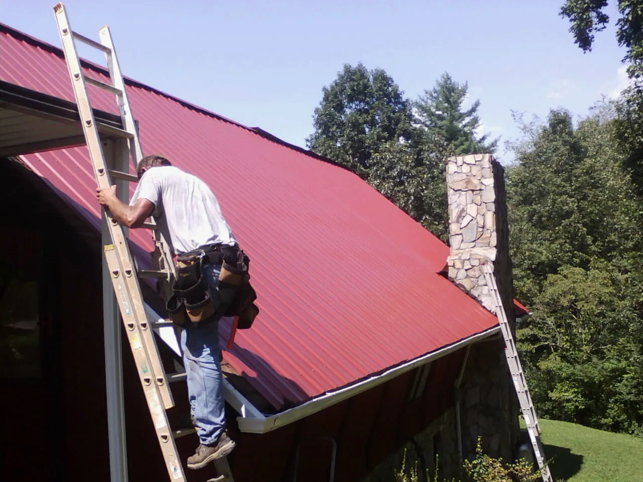 Roofer on a ladder beside a red metal roof and stone chimney — Soffit & Fascia Repair by Roofing Brothers in Raritan